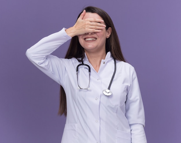 Sonriente joven doctora vistiendo bata médica con estetoscopio cubre los ojos con la mano aislada en la pared púrpura con espacio de copia