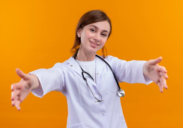 Sonriente joven doctora vistiendo bata médica y estetoscopio estirando las manos en la pared naranja aislada