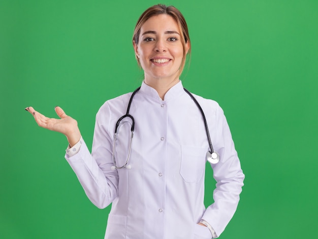 Sonriente joven doctora vistiendo bata médica con estetoscopio extendiendo la mano aislada en la pared verde