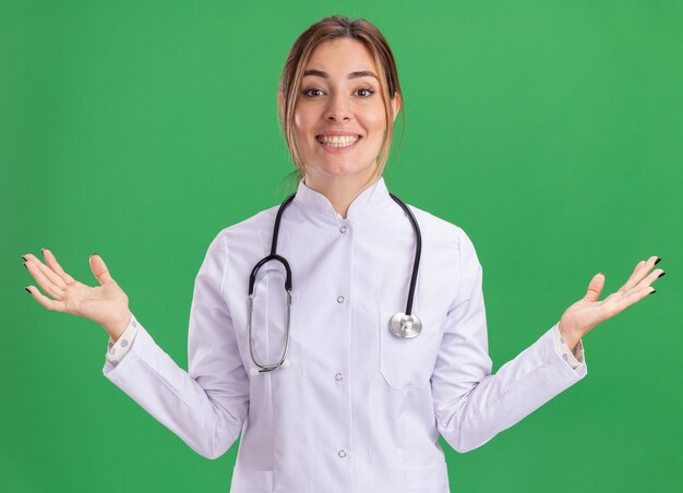 Sonriente joven doctora vistiendo bata médica con estetoscopio extendiendo las manos aisladas en la pared verde