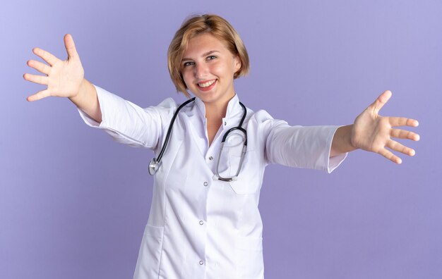 Sonriente joven doctora vistiendo bata médica con estetoscopio extendiendo las manos a la cámara aislada sobre fondo azul.