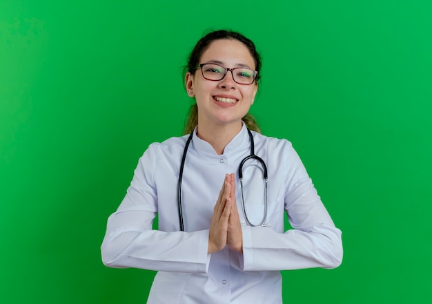 Foto gratuita sonriente joven doctora vistiendo bata médica y estetoscopio y gafas manteniendo las manos juntas aisladas en la pared verde con espacio de copia