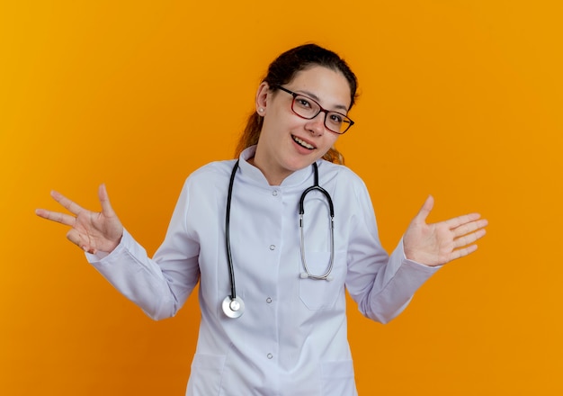 Sonriente joven doctora vistiendo bata médica y un estetoscopio con gafas mostrando diferentes números aislados en la pared naranja