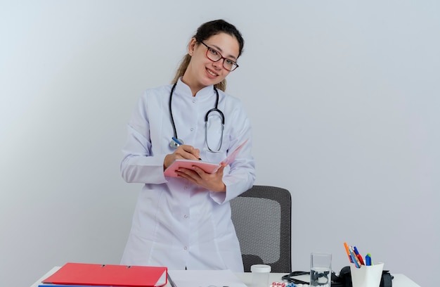 Sonriente joven doctora vistiendo bata médica y estetoscopio y gafas de pie detrás del escritorio con herramientas médicas mirando escribiendo con bolígrafo en el bloc de notas aislado