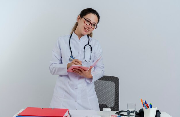 Sonriente joven doctora vistiendo bata médica y estetoscopio y gafas de pie detrás del escritorio con herramientas médicas mirando escribiendo con bolígrafo en el bloc de notas aislado