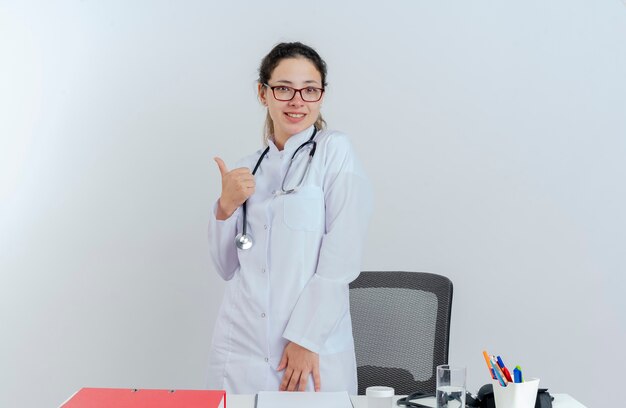 Sonriente joven doctora vistiendo bata médica y estetoscopio y gafas de pie detrás del escritorio con herramientas médicas mirando mostrando pulgar hacia arriba aislado