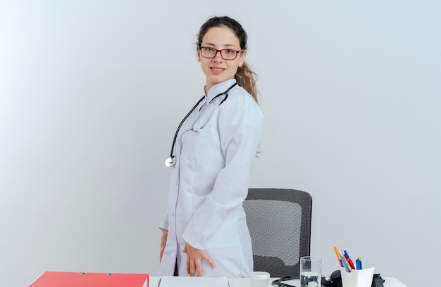 Sonriente joven doctora vistiendo bata médica y estetoscopio y gafas de pie en la vista de perfil detrás del escritorio con herramientas médicas mirando aislado