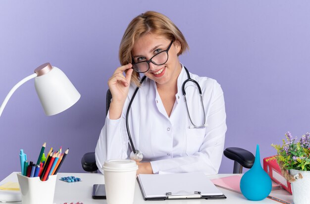 Sonriente joven doctora vistiendo bata médica con estetoscopio y gafas se sienta a la mesa con herramientas médicas aisladas sobre fondo azul.