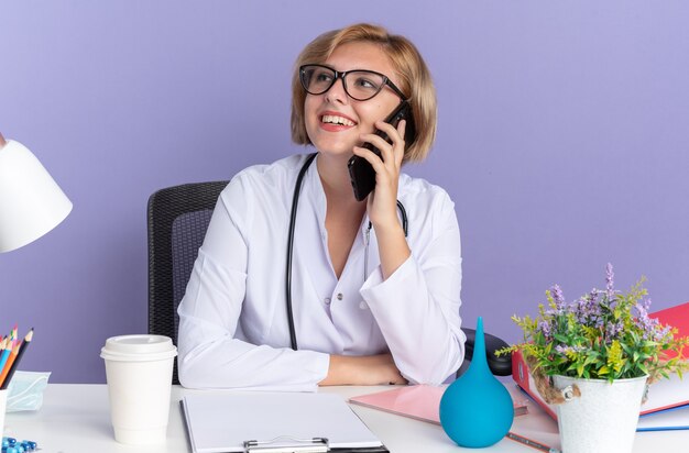 Sonriente joven doctora vistiendo bata médica con estetoscopio y gafas se sienta a la mesa con herramientas médicas habla por teléfono aislado sobre fondo azul.