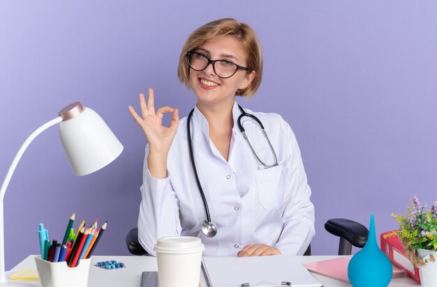 Sonriente joven doctora vistiendo bata médica con estetoscopio y gafas se sienta a la mesa con herramientas médicas mostrando gesto bien aislado sobre fondo azul.
