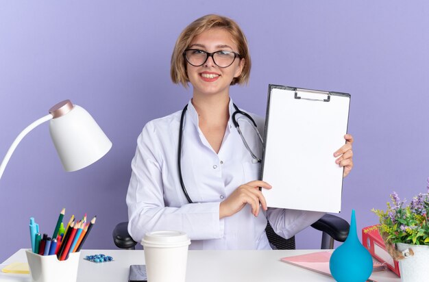 Sonriente joven doctora vistiendo bata médica con estetoscopio y gafas se sienta a la mesa con herramientas médicas sosteniendo el portapapeles aislado sobre fondo azul.