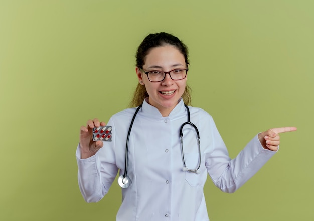 Sonriente joven doctora vistiendo bata médica y estetoscopio con gafas sosteniendo píldoras y puntos en el lado aislado en la pared verde oliva con espacio de copia