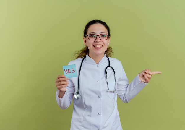 Sonriente joven doctora vistiendo bata médica y estetoscopio con gafas sosteniendo puntos de nota de papel en el lado aislado