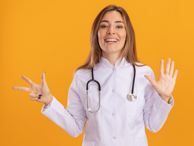 Sonriente joven doctora vistiendo bata médica con estetoscopio mostrando diferentes números aislados en la pared amarilla