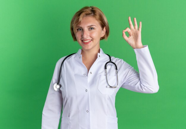 Sonriente joven doctora vistiendo bata médica con estetoscopio mostrando gesto bien aislado sobre fondo verde