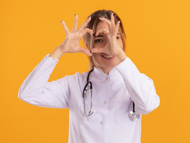 Sonriente joven doctora vistiendo bata médica con estetoscopio mostrando gesto de corazón aislado en la pared amarilla