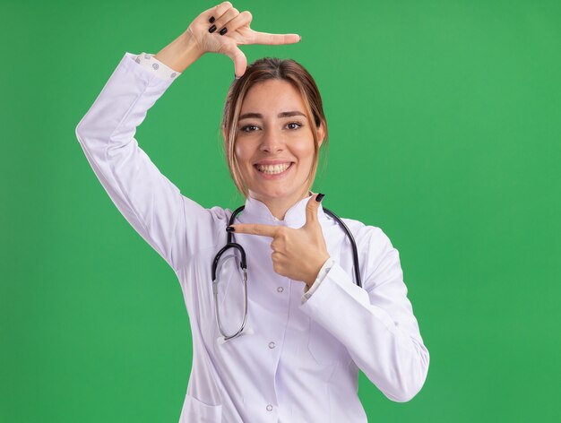 Sonriente joven doctora vistiendo bata médica con estetoscopio mostrando gesto fotográfico aislado en la pared verde