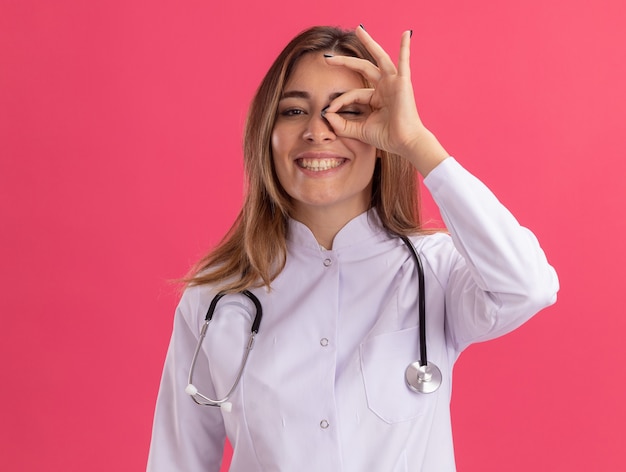 Sonriente joven doctora vistiendo bata médica con estetoscopio mostrando gesto de mirada aislado en la pared rosa