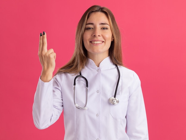 Sonriente joven doctora vistiendo bata médica con estetoscopio mostrando gesto de pistola aislado en la pared rosa