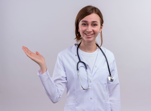 Sonriente joven doctora vistiendo bata médica y estetoscopio y mostrando la mano vacía en la pared blanca aislada