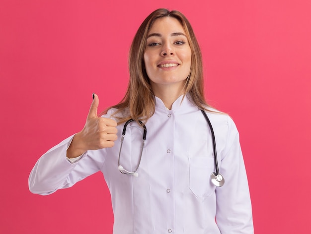 Sonriente joven doctora vistiendo bata médica con estetoscopio mostrando el pulgar hacia arriba aislado en la pared rosa