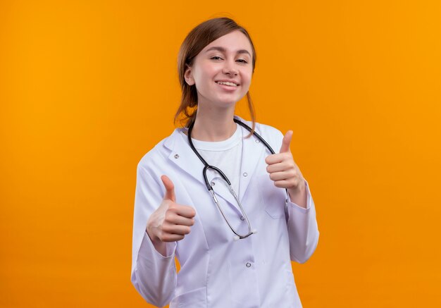 Sonriente joven doctora vistiendo bata médica y estetoscopio mostrando los pulgares hacia arriba en la pared naranja aislada con espacio de copia