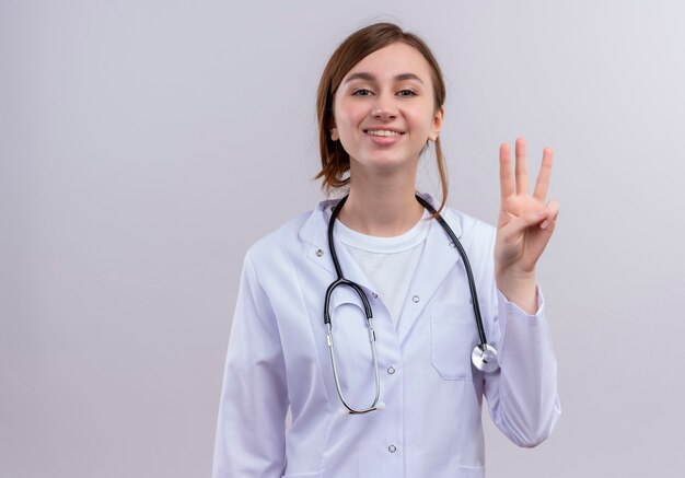 Sonriente joven doctora vistiendo bata médica y estetoscopio mostrando tres en pared blanca aislada con espacio de copia