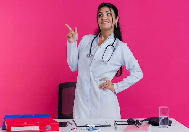 Sonriente joven doctora vistiendo bata médica y un estetoscopio de pie detrás del escritorio con herramientas médicas manteniendo la mano en la cintura mirando y apuntando al lado aislado