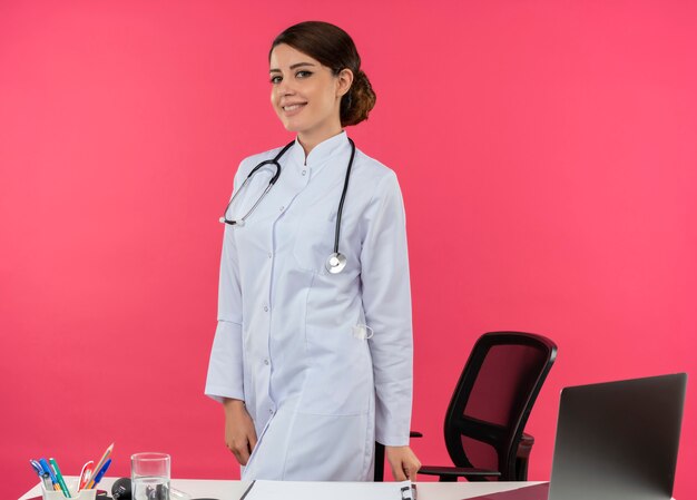 Sonriente joven doctora vistiendo bata médica con estetoscopio de pie detrás de escritorio trabajar en computadora con herramientas médicas con espacio de copia