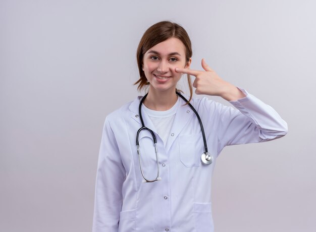 Sonriente joven doctora vistiendo bata médica y estetoscopio poniendo el dedo en la mejilla en la pared blanca aislada con espacio de copia