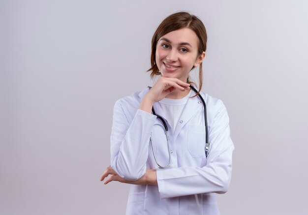 Sonriente joven doctora vistiendo bata médica y estetoscopio poniendo la mano debajo de la barbilla en la pared blanca aislada con espacio de copia