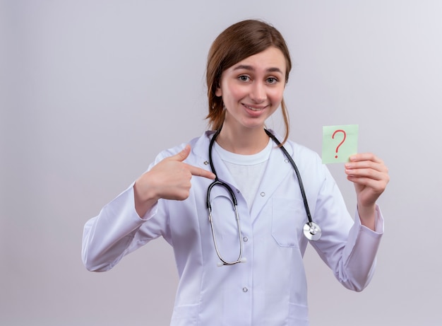 Sonriente joven doctora vistiendo bata médica y estetoscopio y ponting en signo de interrogación escrito en nota de papel en la pared blanca aislada