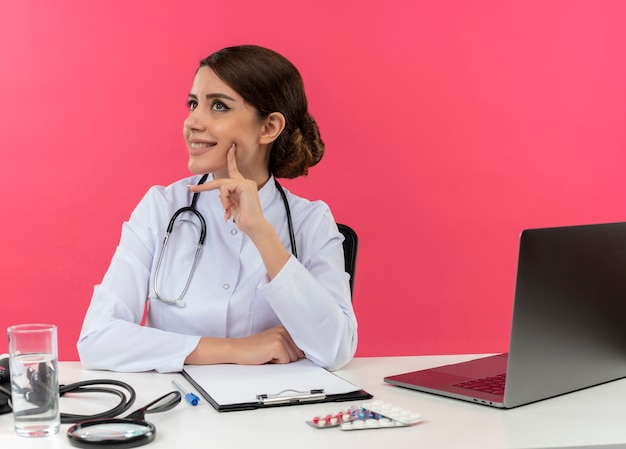 Sonriente joven doctora vistiendo bata médica y estetoscopio sentado en el escritorio con herramientas médicas y portátil girando de cabeza a lado mirando hacia arriba tocando la mejilla con el dedo aislado en la pared rosa