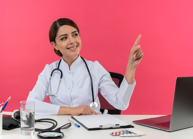 Sonriente joven doctora vistiendo bata médica y estetoscopio sentado en el escritorio con herramientas médicas y portátil mirando y apuntando al lado aislado en la pared rosa