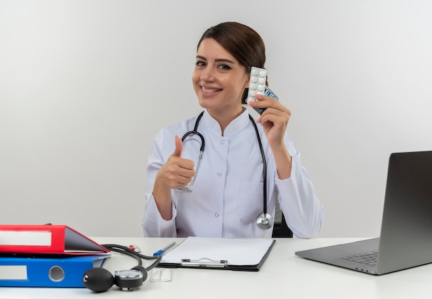 Sonriente joven doctora vistiendo bata médica y estetoscopio sentado en el escritorio con herramientas médicas y portátil sosteniendo medicamentos médicos mostrando el pulgar hacia arriba aislado en la pared blanca