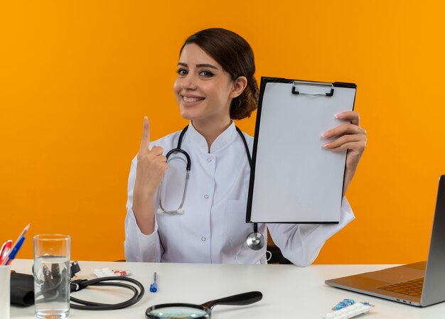 Sonriente joven doctora vistiendo bata médica y estetoscopio sentado en el escritorio con herramientas médicas y portátil sosteniendo portapapeles levantando dedo aislado en pared amarilla