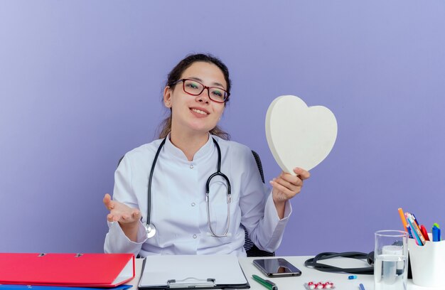 Sonriente joven doctora vistiendo bata médica y estetoscopio sentado en el escritorio con herramientas médicas sosteniendo en forma de corazón mostrando la mano vacía mirando aislado
