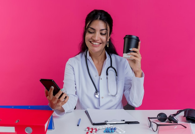 Foto gratuita sonriente joven doctora vistiendo bata médica y un estetoscopio sentado en el escritorio con herramientas médicas sosteniendo una taza de café y un teléfono móvil mirando el teléfono aislado en la pared rosa