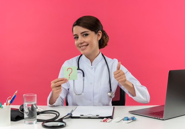 Sonriente joven doctora vistiendo bata médica con estetoscopio sentado en el escritorio trabajar en computadora con herramientas médicas sosteniendo papel de interrogación marque su pulgar hacia arriba en la pared rosa