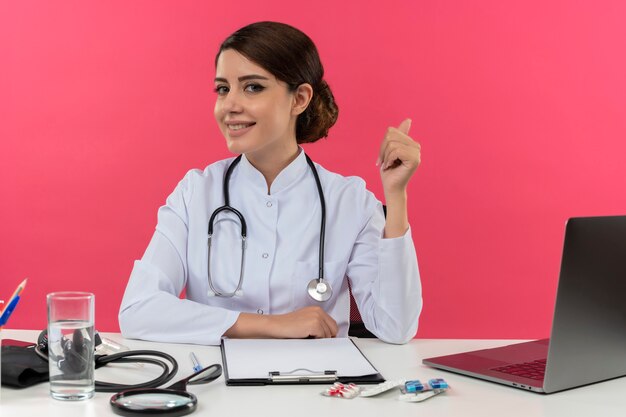 Sonriente joven doctora vistiendo bata médica con estetoscopio sentado en el escritorio trabajar en computadora con herramientas médicas su pulgar hacia arriba con espacio de copia