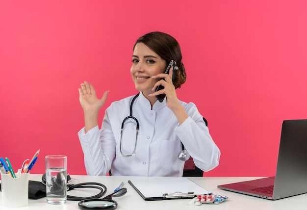 Sonriente joven doctora vistiendo bata médica con estetoscopio sentado en el escritorio, trabajo en computadora con herramientas médicas, habla por teléfono y señala con la mano al lado de la pared rosa