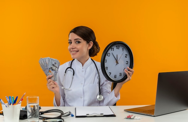 Sonriente joven doctora vistiendo bata médica con estetoscopio sentado en el escritorio de trabajo en computadora con herramientas médicas con reloj de pared y efectivo sobre fondo amarillo de aislamiento