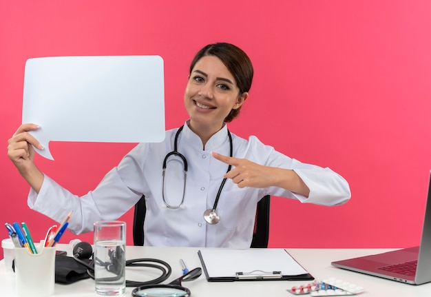 Sonriente joven doctora vistiendo bata médica con estetoscopio sentado en el escritorio, trabajo en computadora con herramientas médicas sosteniendo y apunta a la burbuja de chat en blanco en la pared rosa