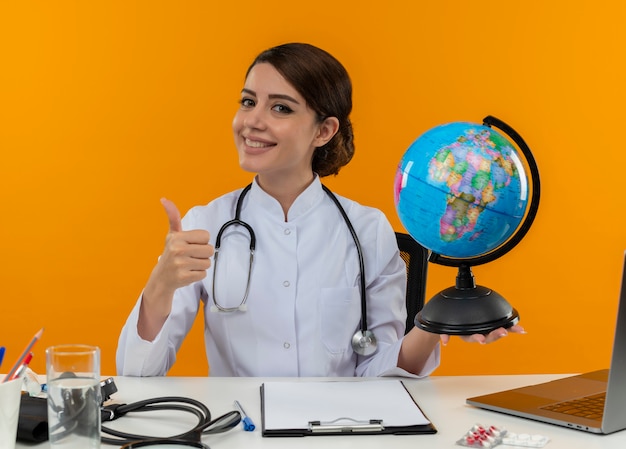 Sonriente joven doctora vistiendo bata médica con estetoscopio sentado en el escritorio de trabajo en computadora con herramientas médicas sosteniendo globo su pulgar hacia arriba sobre fondo amarillo de aislamiento