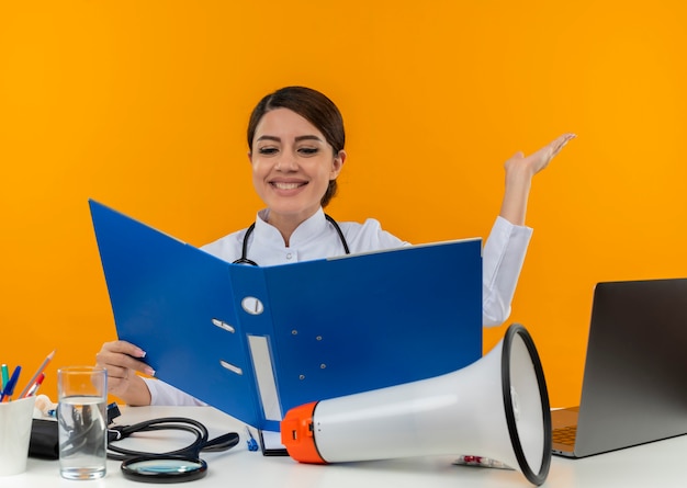 Sonriente joven doctora vistiendo bata médica con estetoscopio sentado en el escritorio de trabajo en la computadora con herramientas médicas sosteniendo y mirando la carpeta y puntos con la mano al lado en la pared amarilla