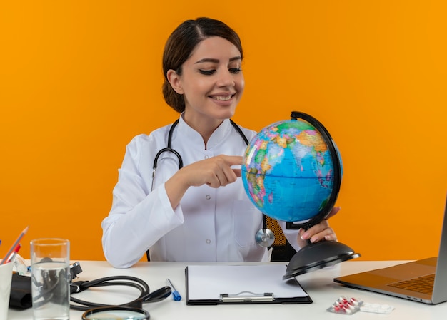 Sonriente joven doctora vistiendo bata médica con estetoscopio sentado en el escritorio de trabajo en la computadora con herramientas médicas sosteniendo y poniendo el dedo en el globo sobre fondo amarillo de aislamiento