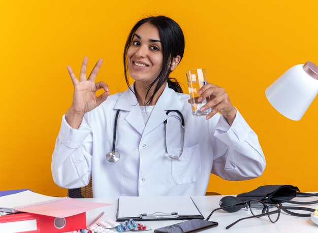 Sonriente joven doctora vistiendo bata médica con estetoscopio se sienta a la mesa con herramientas médicas sosteniendo un vaso de agua mostrando gesto bien aislado sobre fondo amarillo