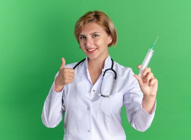 Sonriente joven doctora vistiendo bata médica con estetoscopio sosteniendo la jeringa en la cámara mostrando el pulgar hacia arriba aislado sobre fondo verde