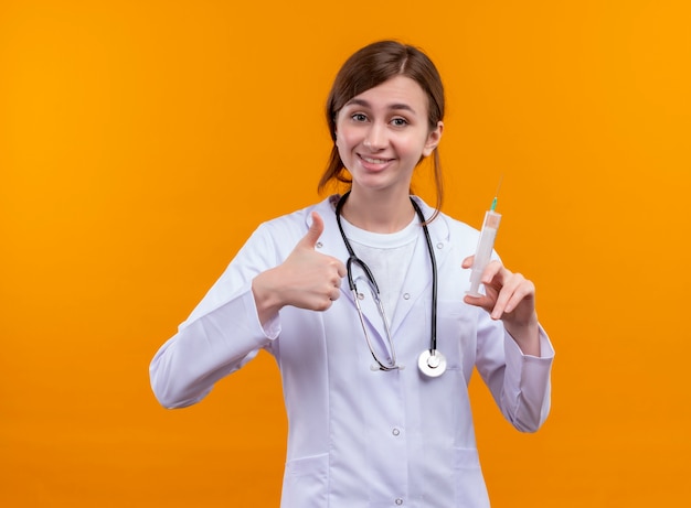 Sonriente joven doctora vistiendo bata médica y estetoscopio sosteniendo la jeringa y mostrando el pulgar hacia arriba en la pared naranja aislada