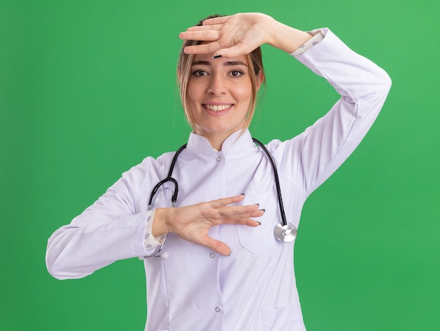 Sonriente joven doctora vistiendo bata médica con estetoscopio sosteniendo la mano en la frente aislada en la pared verde